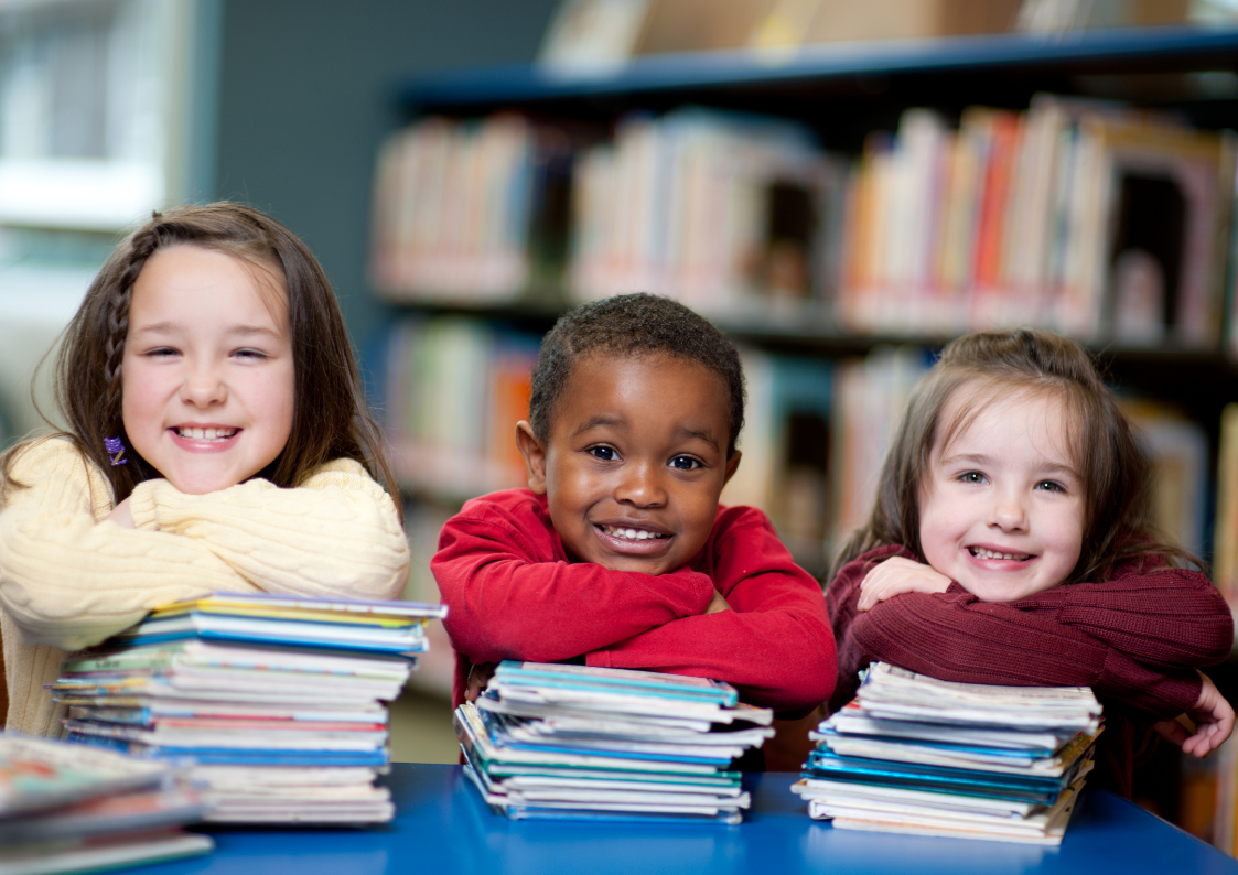 Image of three children with arms crossed leaning on small pile of books with book shelving blurred in the background