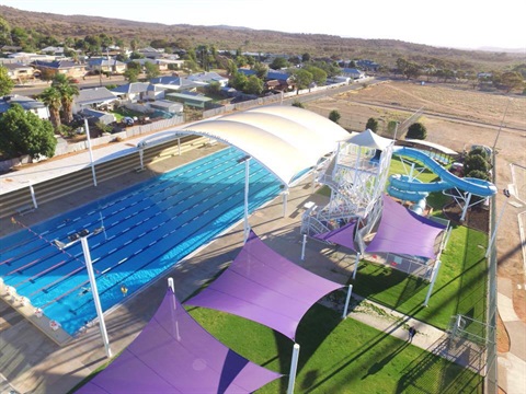 Outdoor fifty meter swimming pool withwhite shade structure at one end and three purple shade sails on grass next to it 